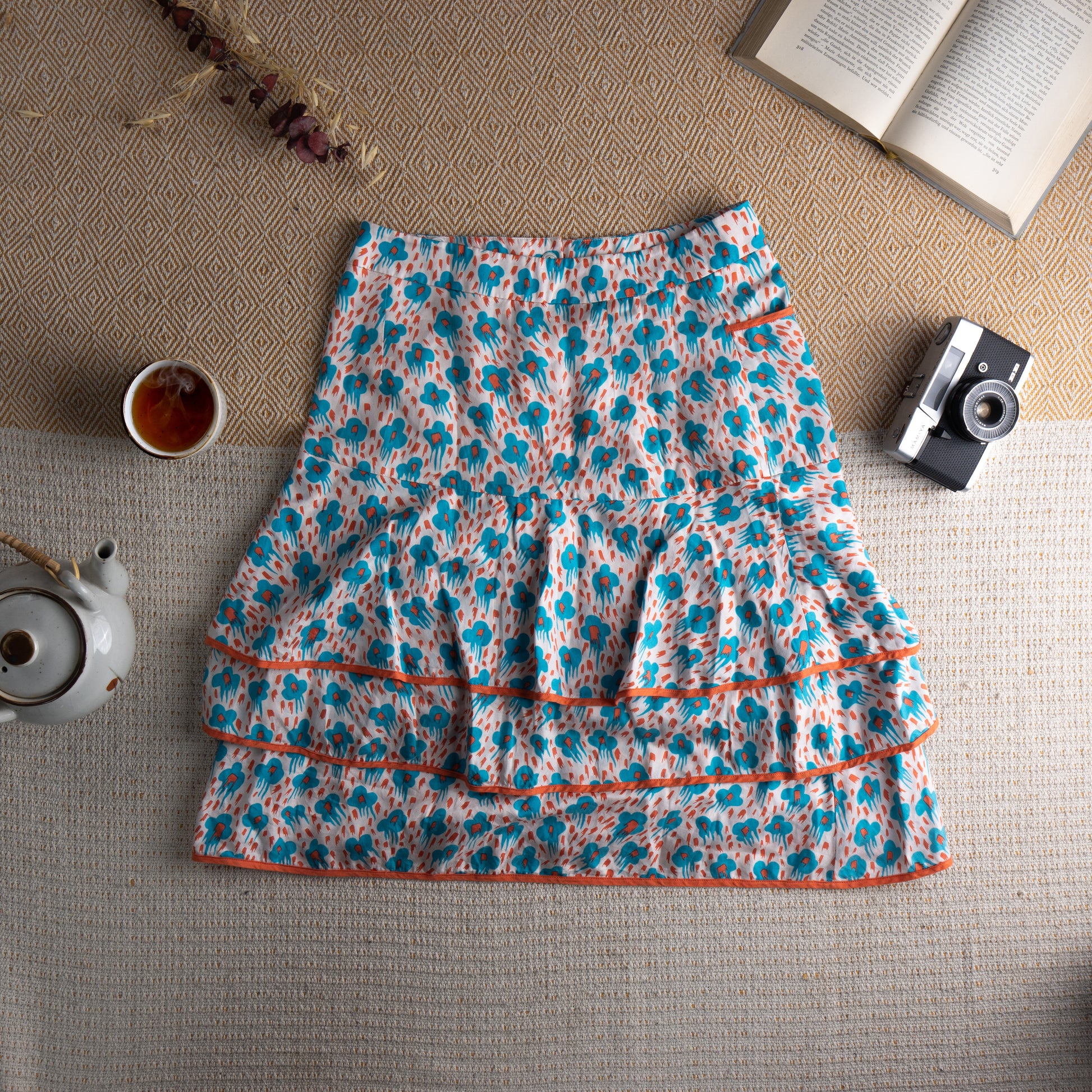 back view Cacharel Archival Deadstock Tiered Ruffle Skirt – white silk-cotton blend with all-over turquoise blue abstract daisy print and red dot scatter, drop waist with three red button front closure, small side pocket with orange trim, double tiered ruffle hem with orange grosgrain ribbon trim, coquette French chic quiet luxury, flat lay editorial shot