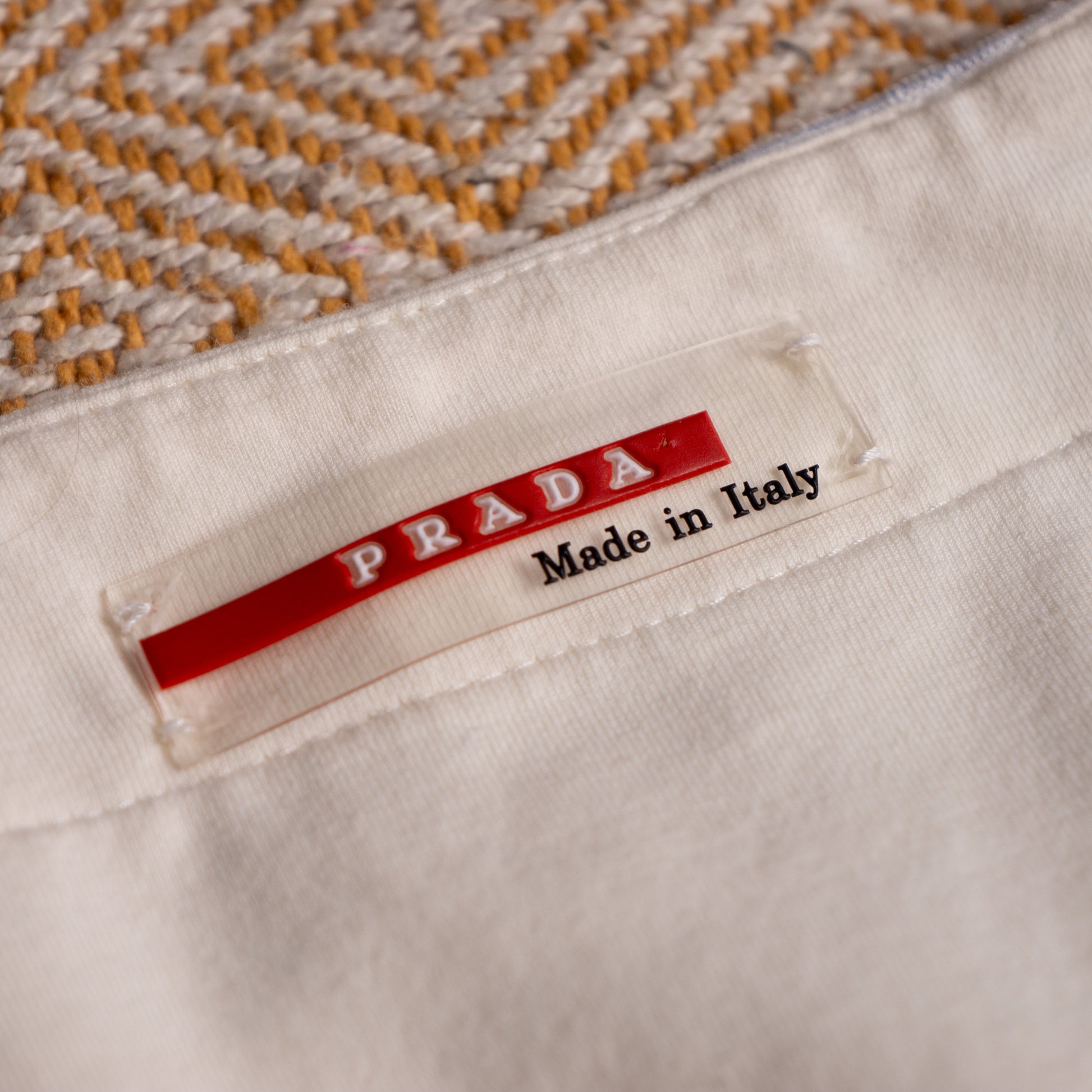 Close-up of a Prada garment tag reading 'PRADA Made in Italy' sewn onto a light cream fabric, with a woven beige and tan textile visible above. Archival Prada Linea Rossa SS2004 Ticker Stripe Cowl Neck Top – ivory cream lightweight cotton, bias-cut diagonal multi-directional ticker stripe print in pale blue and taupe, draped cowl neckline with ruffled edge, sleeveless, empire seam with zip at hem, cropped silhouette, rare red Prada Linea Rossa logo label, IT 46 EU 38 M, product shot on dress form