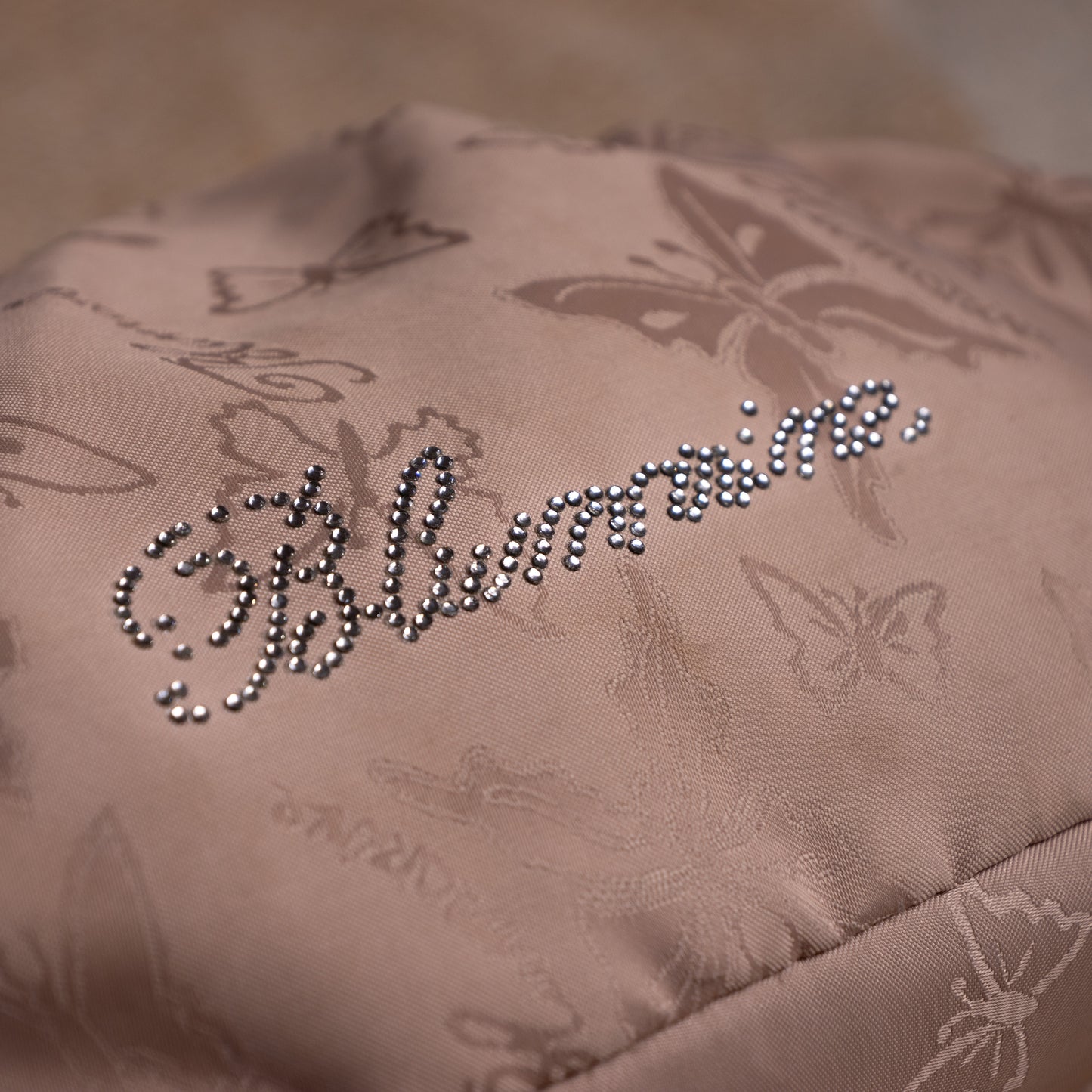 close up Blumarine Y2K Baguette Bag by Anna Molinari – blush beige nylon with tone-on-tone butterfly jacquard print, rhinestone crystal "Blumarine" script logo, dual rolled leather top handles with gold ring hardware, zip closure, Made in Italy, shown on dress form