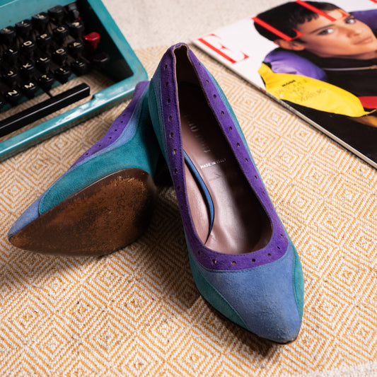 A pair of archival Miu Miu Fall/Winter 2005 Ready-to-Wear runway colorblock pumps. The pointed-toe shoes feature a tripartite design with emerald green suede sides, a cornflower blue suede toe box, and grape purple suede along the throat with classic punched brogue perforations. One shoe stands upright while the other rests on its side, showing the scuffed leather outsole.