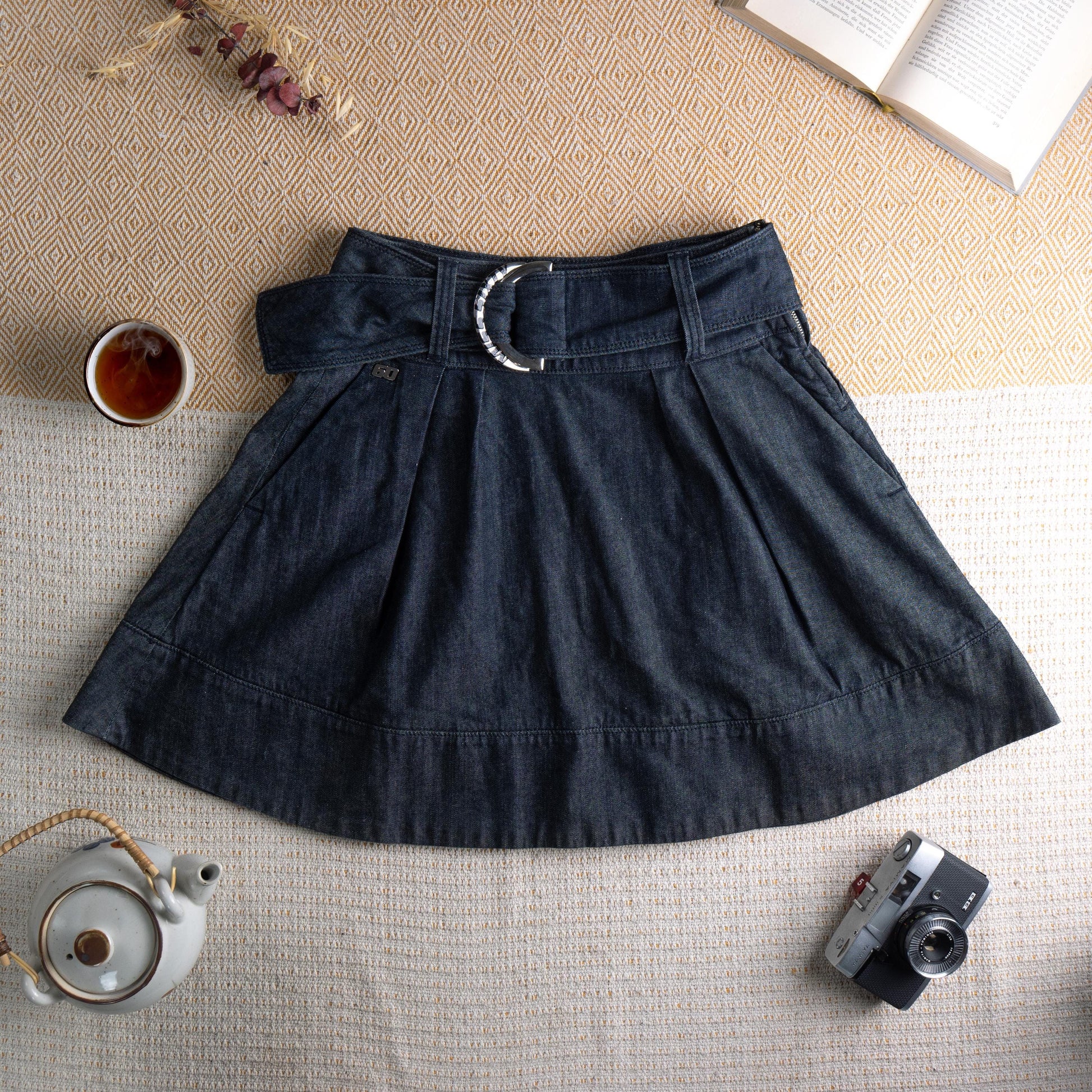 Miss Sixty Y2K Dark Wash Denim Pleated Skater Skirt – deep indigo dark wash denim, high waist with integrated belt and oversized silver Miss Sixty ring buckle, front box pleats, flared skater silhouette, horizontal seam panel at hem, side zip closure, preppy grunge schoolgirl aesthetic, flat lay editorial shot