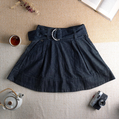 Miss Sixty Y2K Dark Wash Denim Pleated Skater Skirt – deep indigo dark wash denim, high waist with integrated belt and oversized silver Miss Sixty ring buckle, front box pleats, flared skater silhouette, horizontal seam panel at hem, side zip closure, preppy grunge schoolgirl aesthetic, flat lay editorial shot