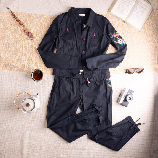 Pristine Deadstock Christian Lacroix Archival Early 2000s Embroidered Cotton Utility Set – jet black crisp cotton, cropped utility jacket with point collar, five dark red enamel logo buttons, two chest flap pockets, fringe-cut hem detail, multicolour floral embroidery patch on left sleeve; matching high-waist cropped tapered trousers with pleated waistband, drawstring tie, fringe-cut hem and matching embroidery at hip, flat lay editorial shot