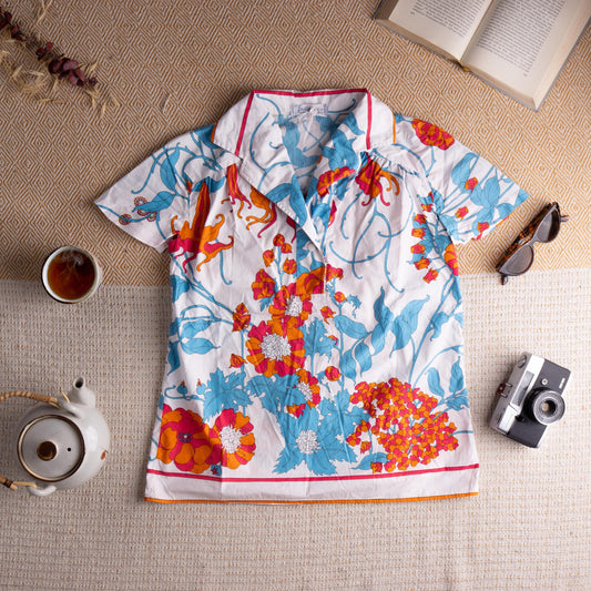 Archival 1970s Emilio Pucci Signed Floral Cotton Blouse – white crisp cotton with large-scale botanical print in turquoise blue, hot pink, orange and white featuring poppies, hydrangeas and scrolling vines, wide point collar with pink contrast trim, short sleeves with gathered shoulder detail, orange contrast hem trim, Pucci signed label, museum quality resort wear, flat lay editorial shot