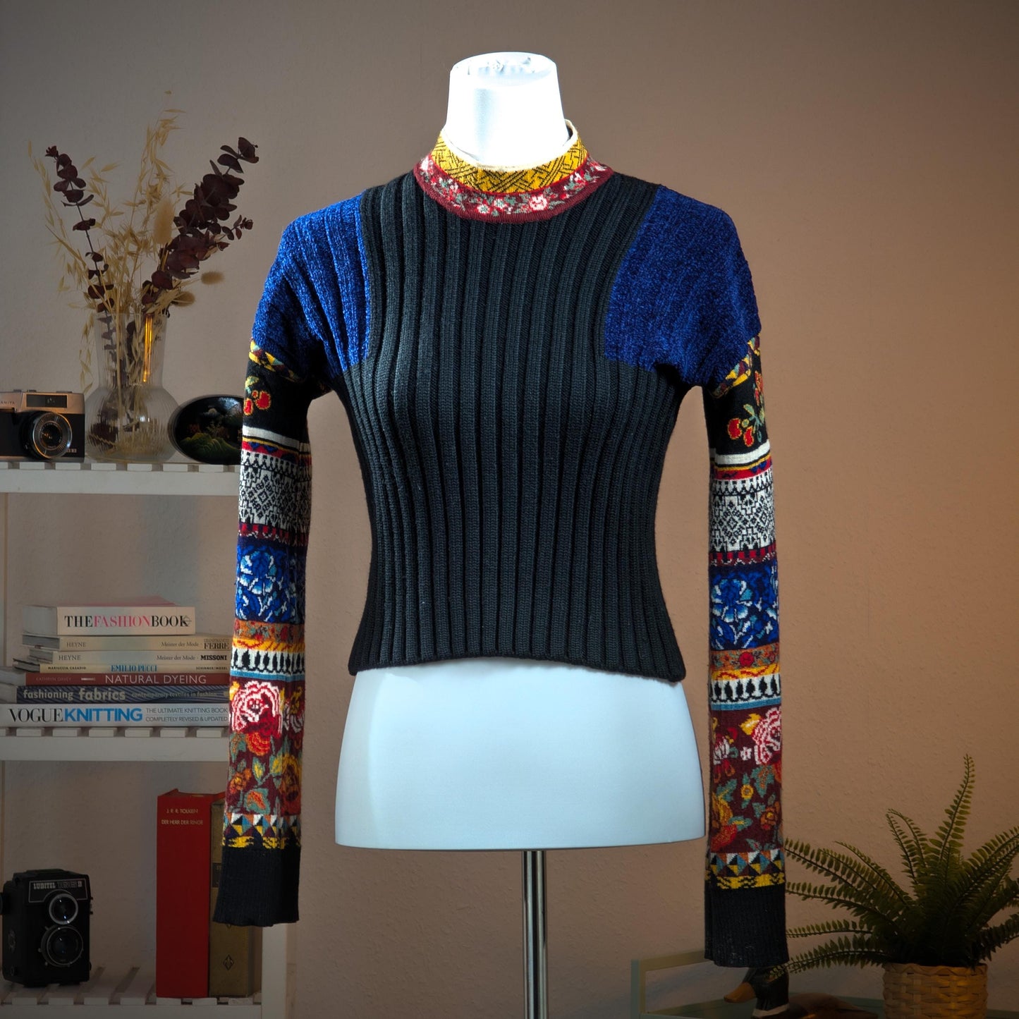 Archival Kenzo Paris 90s Floral Patchwork Ribbed Knit Crop Sweater – charcoal grey wide-rib knit body with cobalt blue lurex pointelle shoulder panels, multicolour patchwork long sleeves featuring folk floral, cherry, geometric diamond and stripe jacquard bands in red, blue, yellow and black, contrast burgundy and yellow folk-print turtleneck collar, boxy cropped silhouette, Takada era, Made in Italy, product shot on dress form