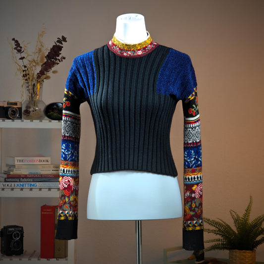 Archival Kenzo Paris 90s Floral Patchwork Ribbed Knit Crop Sweater – charcoal grey wide-rib knit body with cobalt blue lurex pointelle shoulder panels, multicolour patchwork long sleeves featuring folk floral, cherry, geometric diamond and stripe jacquard bands in red, blue, yellow and black, contrast burgundy and yellow folk-print turtleneck collar, boxy cropped silhouette, Takada era, Made in Italy, product shot on dress form