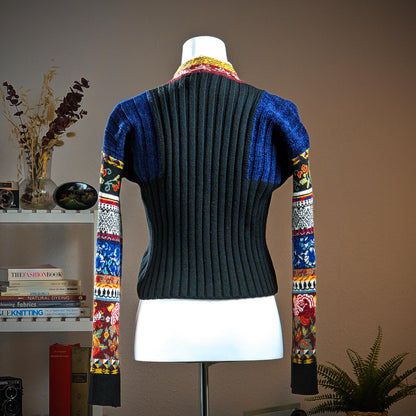back view Archival Kenzo Paris 90s Floral Patchwork Ribbed Knit Crop Sweater – charcoal grey wide-rib knit body with cobalt blue lurex pointelle shoulder panels, multicolour patchwork long sleeves featuring folk floral, cherry, geometric diamond and stripe jacquard bands in red, blue, yellow and black, contrast burgundy and yellow folk-print turtleneck collar, boxy cropped silhouette, Takada era, Made in Italy, product shot on dress form